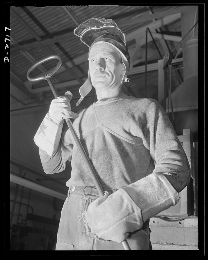 Fiberglass manufacture, Owens-Corning, Toledo, Ohio. In a plant of the Owens-Corning Fiberglass Corporation, a worker stands ready with his iron ladle to transport molten glass from the furnace to the machine that forms it into glass marbles from which it is converted into glass fibers. As one of the men behind the men behind the guns, his work is an essential step in the production of fiberglass yarns being used in a wide variety of military and naval applications