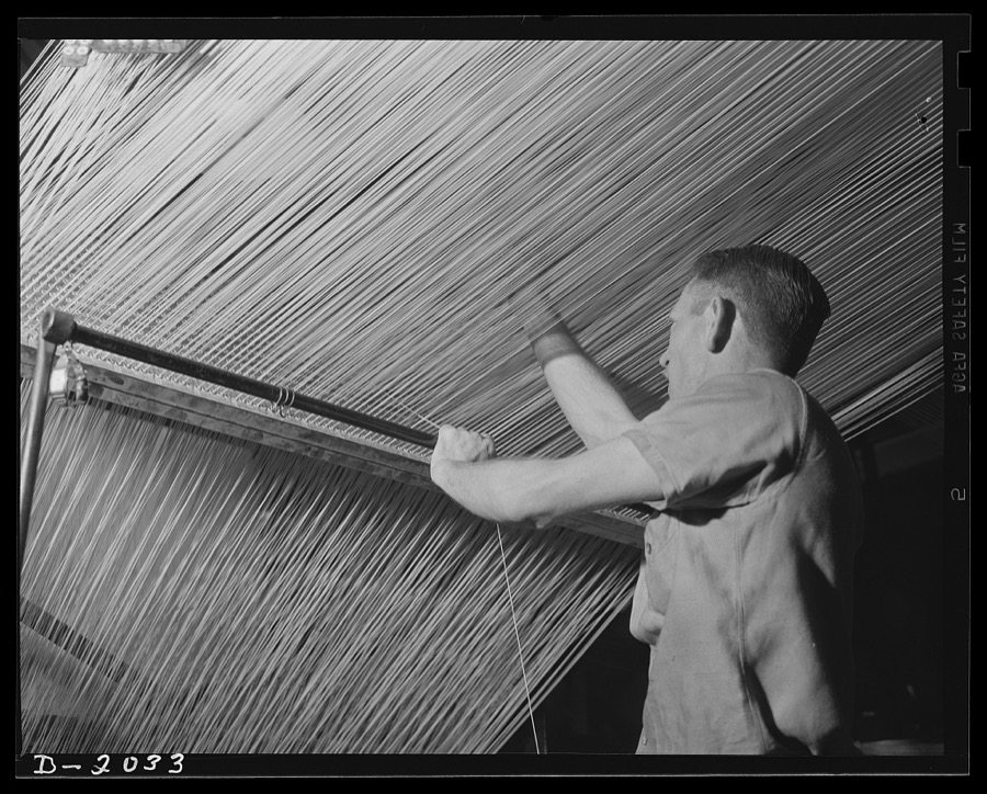 Splicing the line. The loose end in the worker's left hand is feeding an uninterrupted supply of cord to the machine from an emergency spool at the worker's feet. Meantime he searches through the maze of cords to find the one which has broken, to make his splice, and to resume the supply from the regular spool. Firestone (General Tires), Akron, Ohio