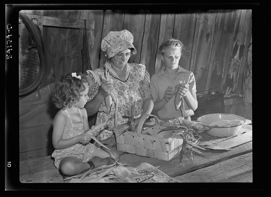 Mrs. Harry Mercer, a young neighbor, and grandchild, husking and removing silk from the corn. The corn will be dried and stored for later use