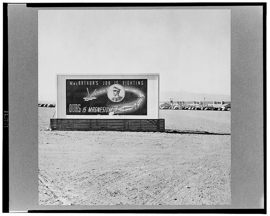 Las Vegas, Nevada. A billboard on the grounds of the gigantic plant of  Basic Magnesium Incorporated, showing tracer bullets and reading "MacArthur's job is fighting. OURS is magnesium." In the background can be seen workers' automobiles and some of the eighty-four foot silos and buildings of the plant