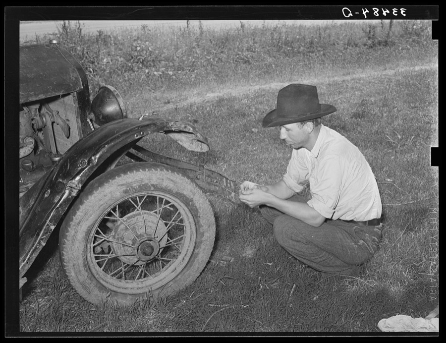 White migrant squatting down in front of his automobile while camped near Prague, Olkahoma. These people use the term "hunkering down" instead of "squatting." "Hunkering down" is a slang term from the Indians