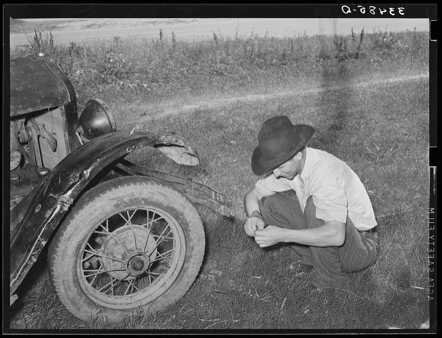 White migrant squatting down in front of his automobile while camped near Prague, Olkahoma. These people use the term "hunkering down" instead of "squatting." "Hunkering down" is a slang term from the Indians