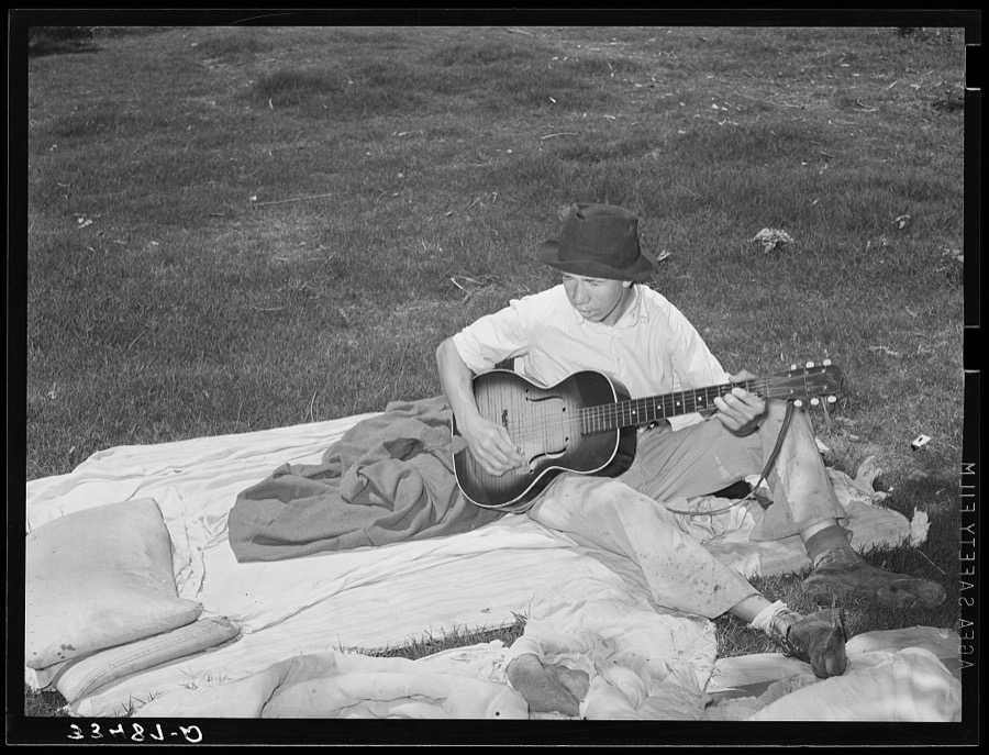 Migrant boy playing the guitar on pallet while camped near Prague, Oklahoma. Lincoln County, Oklahoma