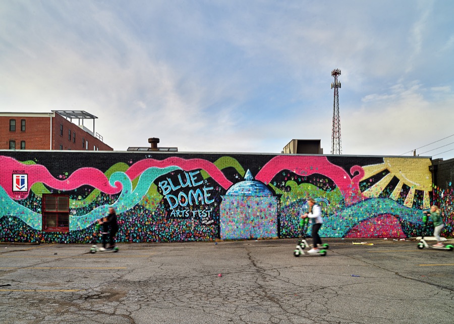 Members of a family or families whiz fast, out of focus to the camera, on scooters in a lot next to a downtown Tulsa, Oklahoma, mural